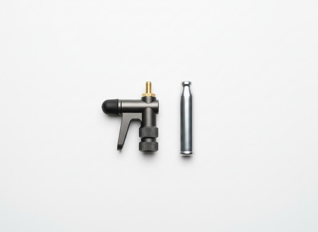 Tête de gonflage Topeak Micro AirBooster ultra-compacte en aluminium usiné posée à côté d'une cartouche de CO2 argentée sur un fond blanc épuré.