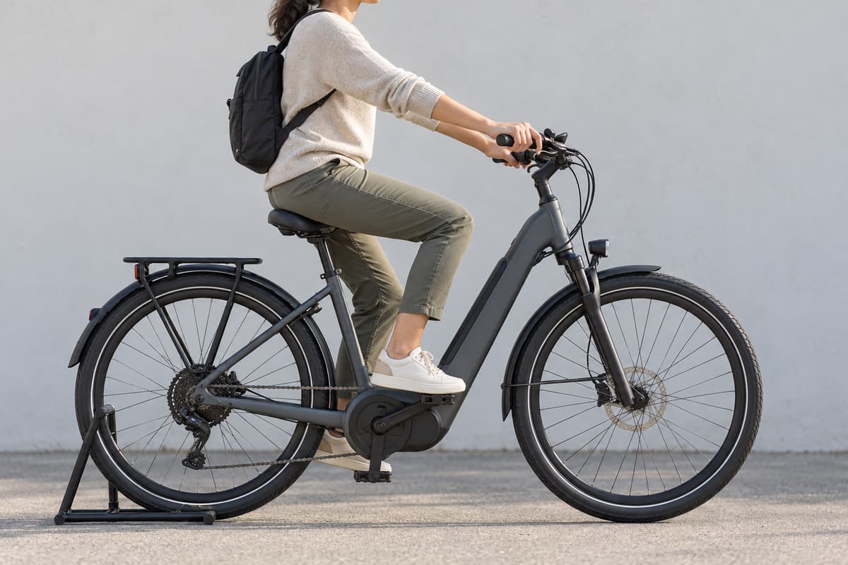 Un adulte de profil assis sur un VTC électrique stabilisé à l’arrêt, les mains sur les poignées et les leviers de frein, un pied sur la pédale basse avec le genou légèrement fléchi, dans une posture détendue.